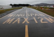 A wet taxiway labeled with the words Wingspan Max 12M at Canberra Airport in August 2020. Taxiway and unserviceability cones appear on the grass next to the taxiway.