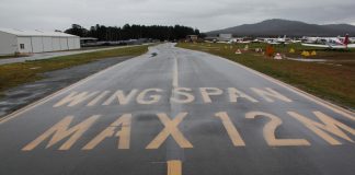 A wet taxiway labeled with the words Wingspan Max 12M at Canberra Airport in August 2020. Taxiway and unserviceability cones appear on the grass next to the taxiway.