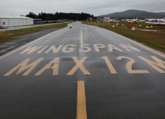 A wet taxiway labeled with the words Wingspan Max 12M at Canberra Airport in August 2020. Taxiway and unserviceability cones appear on the grass next to the taxiway.