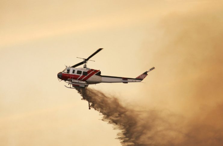Fire fighting helicopter dropping water against a smoky orange sky.