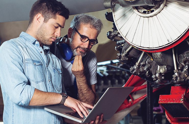 aircraft engineers working on aircraft, referring to and discussing information on a laptop device.