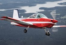 Photo of small red and white aircraft flying with land and water in the background