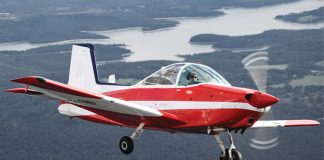 Photo of small red and white aircraft flying with land and water in the background