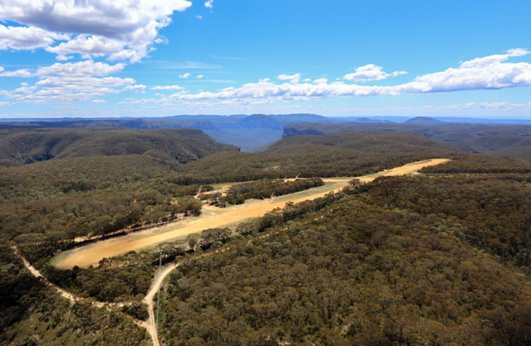 An unsealed runway surrounded by bushland. A blue sky with scattered above.