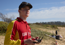 A person wearing a red and yellow top with "UAV Pilot" on the front standing on the beach, holding a remote control and looking up at the sky.