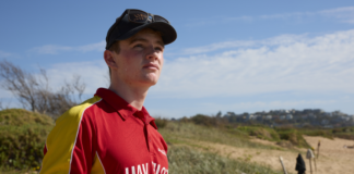 A person wearing a red and yellow top with "UAV Pilot" on the front standing on the beach, holding a remote control and looking up at the sky.
