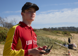 A person wearing a red and yellow top with "UAV Pilot" on the front standing on the beach, holding a remote control and looking up at the sky.