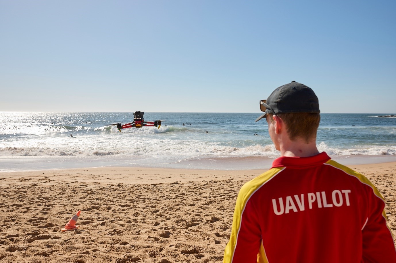 A person wearing a red and yellow top with "UAV Pilot" on the back, stands on a beach, looking at a drone hovering at eye level, with the ocean in the background.