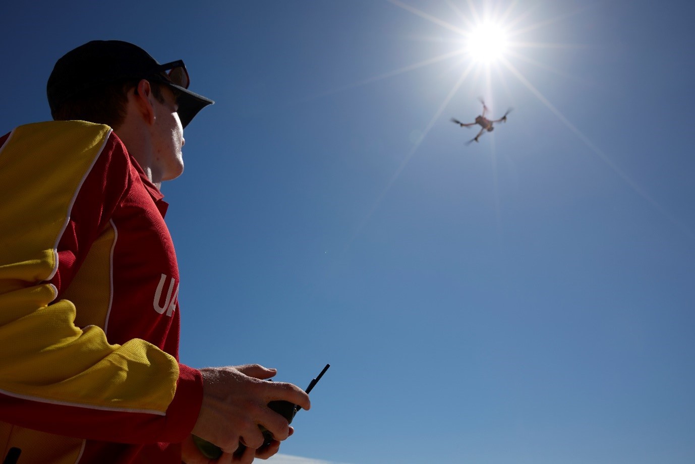 A person holding a remote control looking up at a drone flying high in the sky, against the sun.