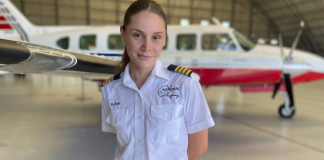 A flight instructor in a white pilot uniform stands in a hangar with a small training aircraft parked behind her.