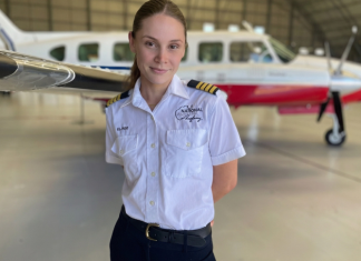 A flight instructor in a white pilot uniform stands in a hangar with a small training aircraft parked behind her.