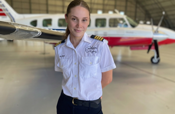 A flight instructor in a white pilot uniform stands in a hangar with a small training aircraft parked behind her.