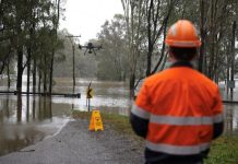 Drone flyer diary: Ben Dawson Photograph shows a flood response scene, with Ben Dawson in high-visibility safety gear observing a drone above submerged roadway and trees. Brown floodwater covers surrounding land, with a warning sign, utility poles, and partially inundated barriers visible.
