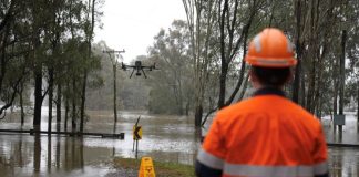 Photograph shows a flood response scene, with Ben Dawson in high-visibility safety gear observing a drone above submerged roadway and trees. Brown floodwater covers surrounding land, with a warning sign, utility poles, and partially inundated barriers visible.