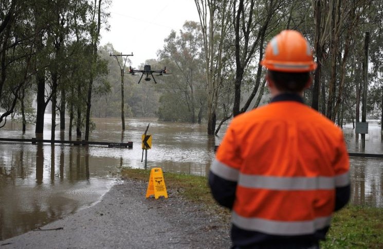 Drone flyer diary - Ben Dawson 1 Photograph shows a flood response scene, with Ben Dawson in high-visibility safety gear observing a drone above submerged roadway and trees. Brown floodwater covers surrounding land, with a warning sign, utility poles, and partially inundated barriers visible.
