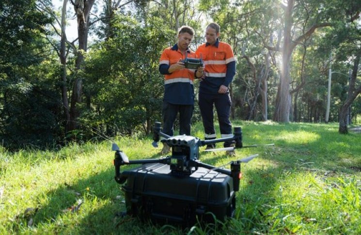 Two people in work uniforms operating a large utility drone in a wooded, grassy area.