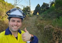Drone flyer diary: Liam Moore Photograph of utility worker in yellow safety jacket and hard hat giving thumbs-up beside a transmission tower. Behind the worker is overgrown vegetation, a second worker, and a sunny outdoor setting.