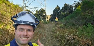 Photograph of utility worker in yellow safety jacket and hard hat giving thumbs-up beside a transmission tower. Behind the worker is overgrown vegetation, a second worker, and a sunny outdoor setting.