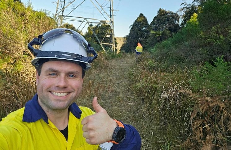 Drone flyer diary - Liam Moore 1 Photograph of utility worker in yellow safety jacket and hard hat giving thumbs-up beside a transmission tower. Behind the worker is overgrown vegetation, a second worker, and a sunny outdoor setting.