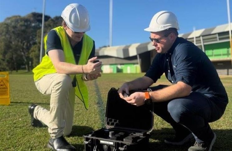 Photograph shows two workers in white hard hats and high-visibility vests examining equipment beside an open protective drone case outdoors.
