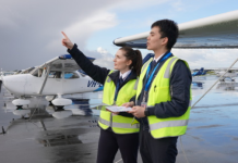 Two pilots in high-visibility vests stand beside a light aircraft on a wet tarmac, looking towards approaching dark clouds.
