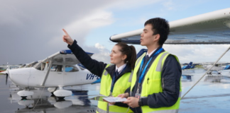 Two pilots in high-visibility vests stand beside a light aircraft on a wet tarmac, looking towards approaching dark clouds.