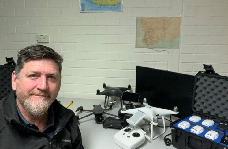 Drone flyer diary - Mathew Hilyard 1 Photograph shows a man beside a white quadcopter drone, remote controller, batteries, monitors, and related equipment.