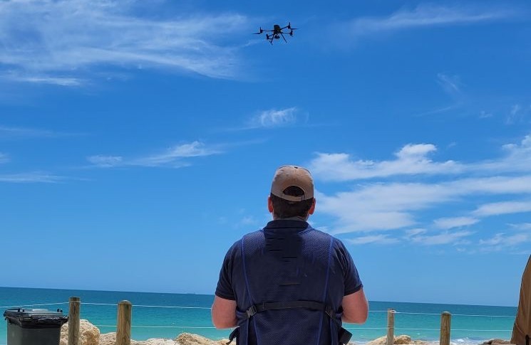 Photograph showing a person piloting a quadcopter above a beach.