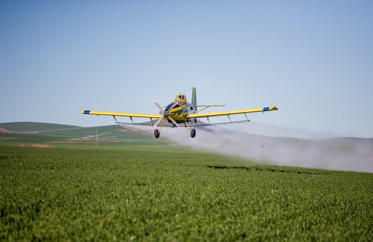 New Toolbox Talks for agricultural aviation safety Yellow agricultural aeroplane flying low over a green crop, spraying a white mist from its wings.